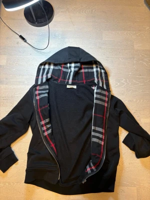 Svart hoodie med rutigt foder - En väldigt snygg och trendig hoddie från burberry! Den är Svart hoodie med dragkedja och huva. Insidan av huvan har ett rutigt mönster i svart, vitt och rött. Broderad detalj på bröstet. Perfekt för en chill och stilren look. Den är M men sitter som S