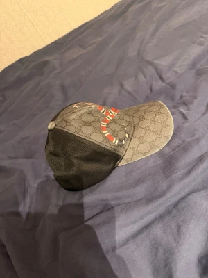 Grå keps med orm och GG-mönster - Snygg keps i grått med klassiskt GG-mönster och en rödvit orm på framsidan. Baksidan är i svart mesh för extra ventilation. Perfekt accessoar för att lyfta din streetwear-look.