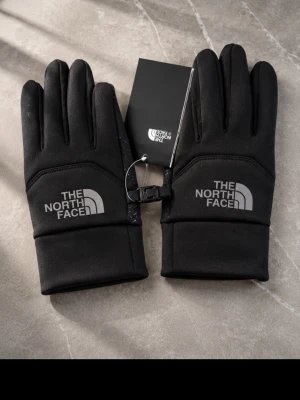 Svarta handskar från The North Face - Svarta handskar från The North Face med logga på ovansidan. Handskarna har en stilren design, justerbart spänne och är tillverkade i ett mjukt syntetmaterial som passar perfekt för kyliga dagar.