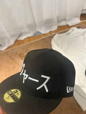 Svart New Era 59FIFTY keps - Säljer en svart New Era 59FIFTY keps med platt skärm och broderad japansk text framtill. Klassisk snapback-modell med New Era-logga på sidan. Kepsen är i ett stadigt material och har en gul etikett på skärmen. Använt ett fåtal gånger, kvitto finns. Endast seriösa köpare!