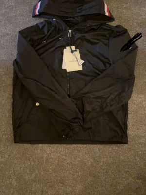 Svart vindjacka med huva - Snygg svart vindjacka med dragkedja och huva. Jackan har vita och röda detaljer på huvan samt praktiska fickor med knappstängning. Tillverkad i ett lätt och vindtätt material, perfekt för blåsiga dagar. Pris kan diskuteras vid snabb affär 
