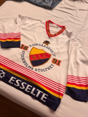 Djurgårdens IF hockeytröja vit - Djurgårdens IF hockeytröja i vitt med klubbens emblem och texten 'Stockholms Stolthet' på bröstet. Tröjan har röda, gula och blå detaljer, randiga ärmar och V-ringad krage. Tillverkad i meshmaterial för bra ventilation. Perfekt för match eller supporterhäng. ifrån 1990-2000 talrt