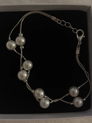 Silverfärgat armband med pärlor - Snyggt armband med dubbla tunna silverfärgade kedjor och stora vita pärlor. Pärlorna är jämnt utspridda längs kedjan och ger en elegant look. Aldrig använt!