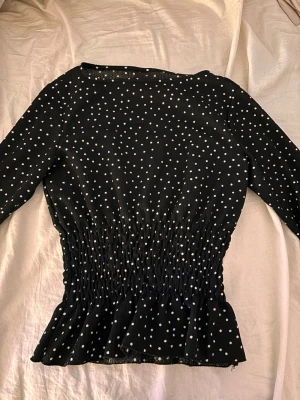 Svart prickig blus från Gina Tricot - Säljer en svart blus från Gina Tricot i storlek XS med vita prickar. Blusen har lång ärm, smockad midja och volang nedtill som ger en snygg siluett. 