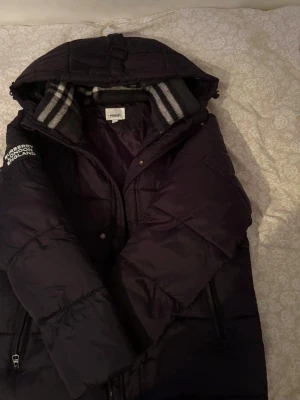 Svart pufferjacka från Burberry - Säljer en svart pufferjacka från Burberry med huva och dragkedja. Jackan har en tydlig Burberry London England-patch på ärmen och randig insida vid kragen. Perfekt för kalla vinterdagar och riktigt snygg streetstil.