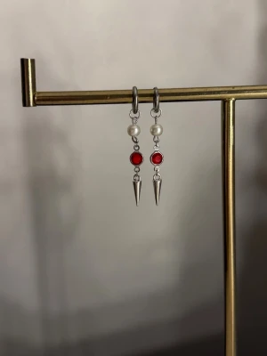 Silvriga örhängen med pärlor och rött - Unika örhängen i silverfärg med vita pärlor, röda stenar och spetsiga metallhängen. Snygg kombination av klassiskt och edgy med pärldetaljer och coola spikes. Perfekta för dig som vill sticka ut med accessoarerna.