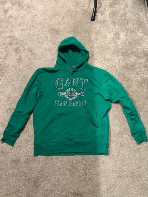 Grön hoodie från GANT New Haven - Snygg grön hoodie från GANT med trycket 'GANT New Haven' och emblem framtill. Klassisk huva och ribbade muddar vid ärmar och nederkant. Perfekt för en avslappnad och sportig stil. Tillverkad i mjukt bomullsmaterial.