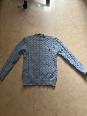 Polo Ralph Lauren tröja  - Ljusblå kabelstickad tröja från Polo Ralph Lauren med rund halsringning och klassisk broderad logga på bröstet. Tröjan har långa ärmar och ribbade muddar vid ärmslut och nederkant. Perfekt för dig som gillar en stilren och tidlös look.