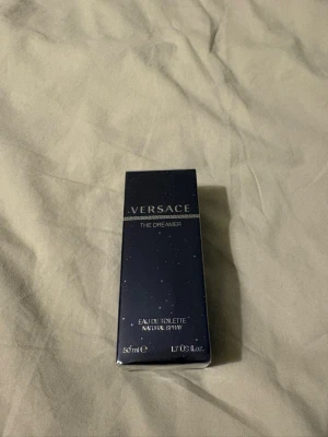 Versace The Dreamer Eau de Toilette - Versace The Dreamer Eau de Toilette i 50 ml flaska. Förpackningen är mörkblå med små vita prickar och har klassisk Versace-design med silverdetaljer. Doften är en ikonisk herrparfym som kombinerar fräscha och kryddiga noter.