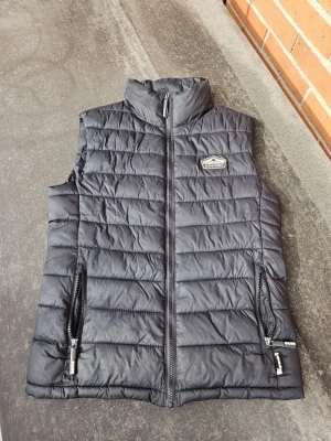 Svart dunväst från Superdry S - Snygg svart dunväst från Superdry med hög krage och två dragkedjefickor framtill. Västen har ett diskret Superdry-märke på bröstet och quiltad design som ger en sportig look. Perfekt att slänga över en hoodie eller tröja för extra värme nu till hösten och vintern. Hör gärna av er om ni undrar något!!!