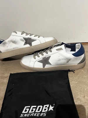Vita GGDB sneakers med grå stjärna - Snygga vita sneakers från Golden Goose med grå stjärna på sidan, beige sula och grå skosnören. Hälpartiet är blått och skon har en cool, lite sliten look. Tillverkade i läder och har GGDB/SSTAR-tryck på sidan.