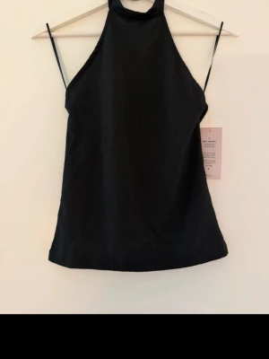 Svart ribbad halterneck topp - Snygg svart ribbad halterneck topp med tunna axelband och hög hals. Toppen har en figurnära passform och är tillverkad i ett stretchigt material som ger en bekväm känsla. Perfekt att styla med jeans eller kjol för en trendig look. 