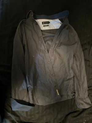 Mörkblå skjorta från Riley - Snygg mörkblå skjorta från Riley med diskreta prickar på insidan av kragen och kontrastfärgad innerkrage. Skjortan har knappar framtill och lång ärm, perfekt för en stilren look.