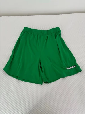 Gröna shorts från Kappa - Gröna sportiga shorts från Kappa med elastisk midja och logga på vänster ben. Perfekta för träning eller chill dagar. Tillverkade i ett lätt och svalt syntetmaterial som andas bra.