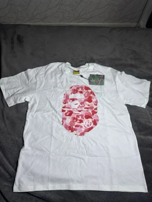 Bape t-shirt - Säljer en vit t-shirt med ett stort tryck av ett rosa och rött camomönster i form av ett ansikte både fram och bak. T-shirten har korta ärmar och en klassisk passform. Perfekt för dig som gillar streetwear och vill sticka ut.
