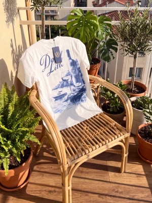 Vit t-shirt med blått tryck - Vit t-shirt med stort blått tryck på ryggen som visar en vinflaska, glas och strandmotiv. T-shirten har klassisk passform och korta ärmar, perfekt för dig som gillar stilrena och kreativa prints.