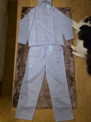 Nocta Nike loungewear-set -  Passar som (L till Xl) Säljer ett ljusblått loungewear-set från Nike med hoodie och matchande byxor. Setet har dragkedjor på fickorna och diskreta Nike-loggor på både överdel och byxa. Materialet ser mjukt och bekvämt ut, perfekt för chill dagar hemma.