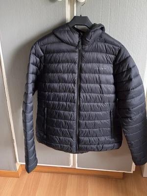 Svart dunjacka från Ocean Works - Snygg svart dunjacka från Ocean Works med huva och dragkedja framtill. Jackan har två sidofickor och quiltad design som ger en modern look. Perfekt för kalla vinterdagar och enkel att matcha med olika outfits.