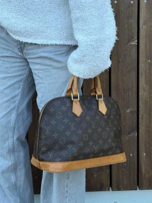 Louis Vuitton brun monogram handväska - Klassisk handväska från Louis Vuitton i brunt monogrammönstrat canvas med detaljer i ljust läder. Väskan har två handtag och rundad form, perfekt för dig som gillar tidlös och lyxig stil. Ikonisk LV-logga över hela väskan och snygga läderdetaljer.