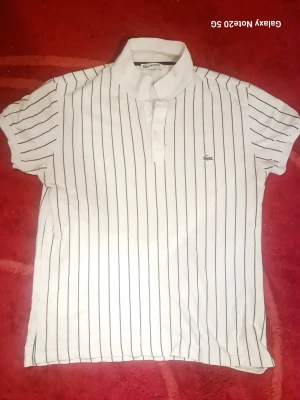 Vit randig pikétröja från Lacoste - Vit pikétröja från Lacoste med svarta vertikala ränder och klassisk krage. Tröjan har korta ärmar och knäppning framtill. Ikoniska krokodilloggan på bröstet. Perfekt för en sportig och stilren look.