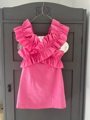 Rosa volangklänning från Zara - Supersöt rosa kortklänning från Zara med stora volanger över axlarna och bröstet. Klänningen har en figurnära passform och är ärmlös, perfekt för dig som vill sticka ut med en färgglad och trendig look.