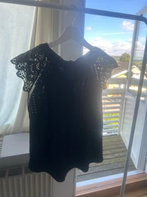 Svart spetsdetaljerad topp med knyt - Säljer en svart topp med fina spetsdetaljer på ärmarna och knyt i nacken. Toppen har en luftig passform och är tillverkad i ett lätt material som känns bekvämt. Perfekt för dig som gillar romantiska detaljer och vill ha något extra till din outfit.