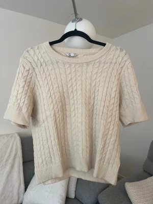 Beige kabelstickad tröja kort ärm - Kabelstickad tröja i beige med korta ärmar och rund hals. Tröjan har ribbade muddar vid ärmslut och nederkant, vilket ger en klassisk och stilren look. Perfekt för lager på lager och enkel att matcha med olika outfits.