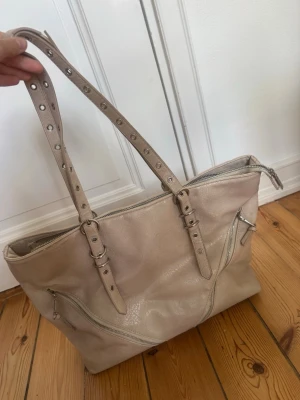 Beige shoppingväska med detaljer - Stilren beige shoppingväska i konstläder med justerbara axelband och silverfärgade dragkedjor framtill. Väskan har en rymlig insida och coola bältesdetaljer på handtagen. Perfekt för dig som vill ha plats för allt du behöver under dagen. Defekter såsom slitningar på ”lädret” och även slitning vid axelbandet, dock går det säkert lätt att sy igen. 