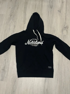 Svart hoodie The Great Norrland - Snygg svart hoodie med vit text 'The Great Norrland' på bröstet och vit dragsko i huvan. Klassisk passform med lång ärm och ribbade muddar. Perfekt för dig som gillar streetwear och vill ha något bekvämt och stilrent. 