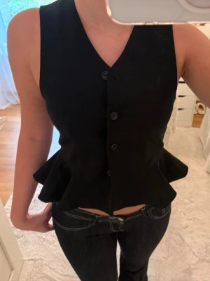 Svart väst med volangkant - Säljer en svart väst med v-ringning och knappar framtill. Västen har en snygg volangkant nedtill som ger en cool och trendig look. Perfekt att styla över en t-shirt eller skjorta för en extra touch.