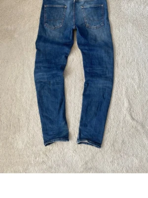 Blå jeans från Jack & Jones - Säljer ett par klassiska blå jeans från Jack & Jones med fem fickor och snygga slitningar. Jeansen har normal midja, raka ben och är gjorda i mjukt denimtyg. Perfekta för en avslappnad och trendig look.