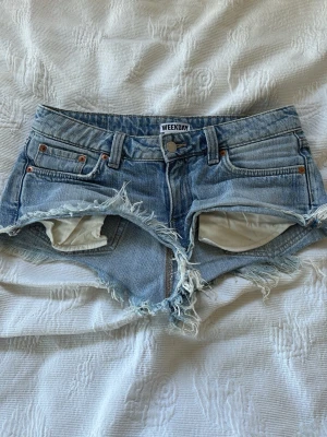 Blå jeansshorts från Weekday - Säljer ett par blå jeansshorts från Weekday med råa, fransiga kanter och klassisk femficksdesign. Shortsen har låg midja och slitna detaljer för en avslappnad look. Perfekta för varma dagar och festivaler.