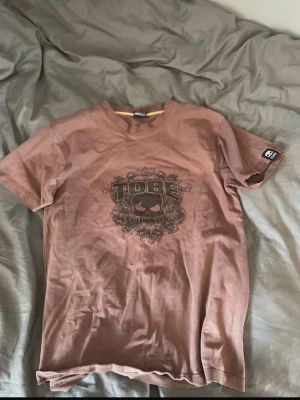 Brun Tobe t-shirt med tryck - Säljer en brun t-shirt från Tobe med ett stort svart tryck på bröstet som föreställer en döskalle och texten 'TOBE TECHNOLOGY'. T-shirten har rund hals och korta ärmar. Perfekt för dig som gillar streetwear och vill ha något unikt i garderoben.
