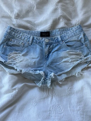 Ljusblå slitna jeansshorts - Säljer ett par ljusblå jeansshorts med fransig kant och slitna detaljer framtill. Shortsen har klassisk femficksmodell, knappgylf och bälteshällor. Perfekta för sommaren och ger en avslappnad vibe.