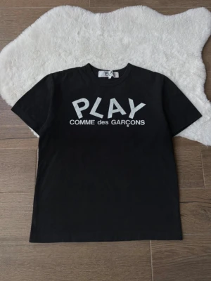 Svart PLAY Comme des Garçons t-shirt - Säljer en svart t-shirt från Comme des Garçons PLAY med vit logga och text framtill. Klassisk rak passform och rund halsringning. Perfekt för dig som gillar stilrena och ikoniska plagg.