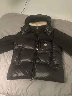 Svart dunjacka  - Snygg svart dunjacka med glansig finish, stor huva och mjukt foder i kragen. Jackan har en dragkedja framtill och quiltad design. Perfekt för kalla vinterdagar och riktigt varm.