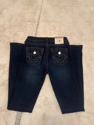 True Religion Jeans - Snygga mörkblå jeans från True Religion med klassiska kontrastsömmar och ikoniska fickdetaljer bak. Jeansen har raka ben och är tillverkade i denim med en lätt stretch. Fickorna har vita knappar och dekorativa sömmar som ger en unik look.