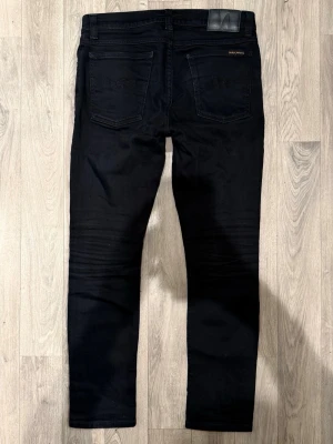 Nudie Jeans Tilted Tor Svarta W31/L30 - Slim Fit - Nudie Jeans Tilted Tor ”Dry Ever Black” svarta jeans med slim fit-passform, storlek W31/L30, midja 40 cm, ytterbenslängd 98 cm, benöppning 15,5 cm, modellen är 185cm & väger 65kg. Det finns ett hål i vänster ficka. I övrigt i fint skick.