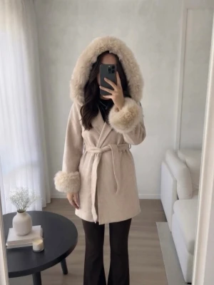 Pälskappa med knytskärp  - Säljer en elegant beige kappa med äkta päls på huva och ärmslut. Kappan har en knytning i midjan och är perfekt för kyligare dagar. Den har en mjuk och lyxig känsla, vilket gör den till ett stilrent val för höst och vinter. S/M