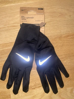 Nike Pacer handskar  - Säljer ett par svarta Nike Pacer Lightweight löparhandskar med reflekterande Swoosh-logga på ovansidan. Handskarna är tunna och smidiga, perfekta för löpning och har touch-kompatibla fingertoppar. Tillverkade i ett mjukt och stretchigt material.                             Size:L