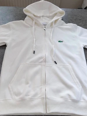 Vit zip hoodie från Lacoste - Säljer en clean vit hoodie från Lacoste med dragkedja, huva med dragsko och klassisk krokodil-logga på bröstet. Hoodien har känguruficka framtill och ribbade muddar. Perfekt för dig som gillar stilren och sportig look.