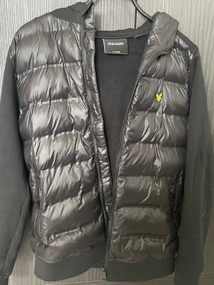 Svart pufferjacka från Lyle & Scott - Snygg svart pufferjacka från Lyle & Scott med quiltad framsida och huva. Jackan har dragkedja, mjuka ärmar i sweatshirtmaterial och den klassiska gula loggan på bröstet. Perfekt för kalla dagar och riktigt skön att bära.