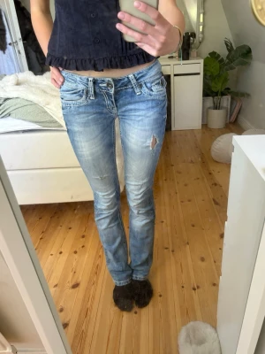 Ljusblå bootcut jeans med slitning - Säljer ett par ljusblå bootcut jeans med slitningar och snygga detaljer på bakfickorna. Jeansen har låg midja, klassisk femficksmodell och ett litet hål på ena låret. Perfekta för en avslappnad och trendig look. Jeansen är lagade på baksidan och passar mig som brukar ha 25/32.