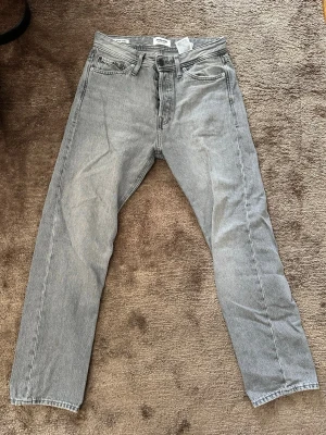 Grå raka jeans i klassisk modell - Säljer ett par grå jeans med rak passform och fem fickor. Jeansen har en klassisk look med normal midja och dragkedja framtill. Materialet är denim i 100% bomull och färgen är ljusgrå, perfekt till en avslappnad stil.