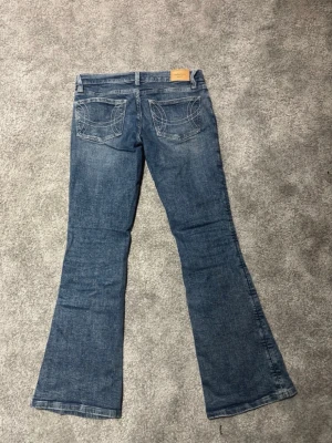Blå bootcut jeans med broderade fickor - Säljer ett par blå bootcut jeans med snyggt slitna detaljer och broderade bakfickor. Jeansen har klassisk femficksmodell och normal midja. Perfekta för dig som gillar en avslappnad men trendig look. Nästan helt oanvända!