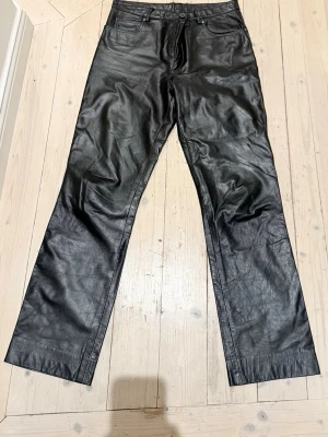 Svarta raka byxor i skinn - Säljer ett par svarta byxor i skinn med rak passform och klassisk femficksdesign. Byxorna har hög midja och stängs med knapp och dragkedja. Perfekta för en edgy och trendig look.