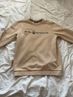 Beige sweatshirt från Sail Racing - fraktar idag skit billigt Snygg beige sweatshirt från Sail Racing med svart logga tryckt på bröstet. Tröjan har rund halsringning, långa ärmar och mjukt, borstat innerfoder för extra komfort. Perfekt för en avslappnad och stilren look.