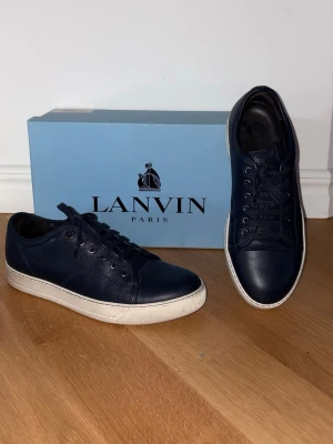 Mörkblå Lanvin sneakers i läder - Snygga mörkblå sneakers från Lanvin i slätt och texturerat läder. Skorna har klassisk låg profil, vita sulor och matchande mörkblå skosnören. Perfekta för dig som vill ha en stilren och exklusiv look.