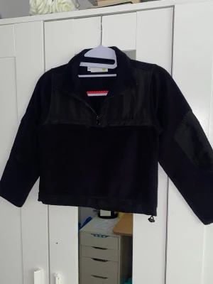 Svart fleece tröja med dragkedja - En svart fleece tröja med halv dragkedja framtill och hög krage. Tröjan har förstärkta partier på axlar och armar samt justerbar nederkant med snörning. Perfekt för kalla dagar och enkel att matcha med jeans eller joggers.