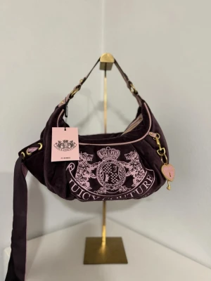 Mörklila axelväska Juicy Couture - Snygg mörklila axelväska i mjuk velour från Juicy Couture med rosa detaljer och broderad logga framtill. Väskan har guldfärgade metalldetaljer, justerbar axelrem och ett hjärtformat hänge. Perfekt för dig som gillar en trendig och lyxig look.